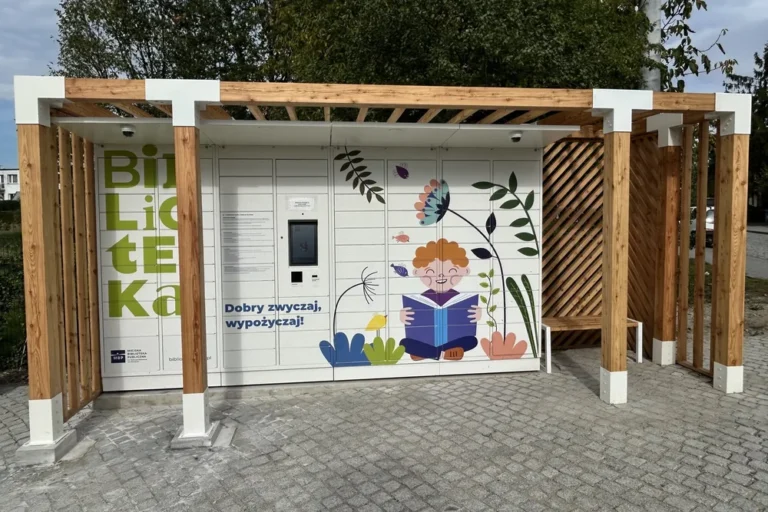 Drewniana pergola z białymi stalowymi detalami wokół książkomatu i ławką po prawej stronie