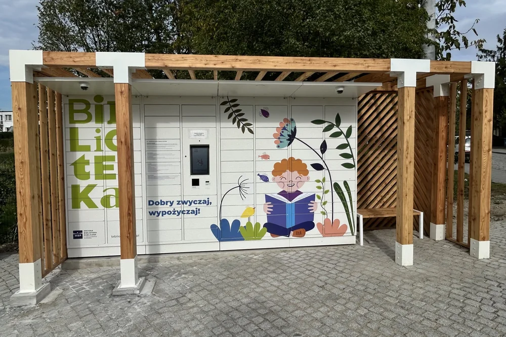 Drewniana pergola z białymi stalowymi detalami wokół książkomatu i ławką po prawej stronie