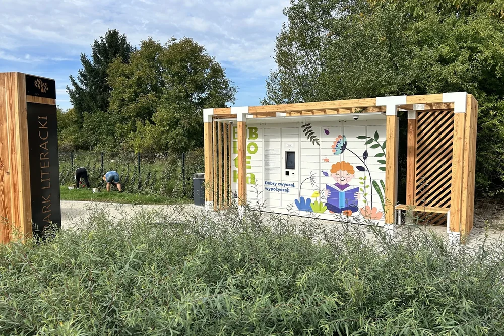 Pergola z książkomatem wśród zieleni, obok drewnianego oznaczenia Parku Literackiego